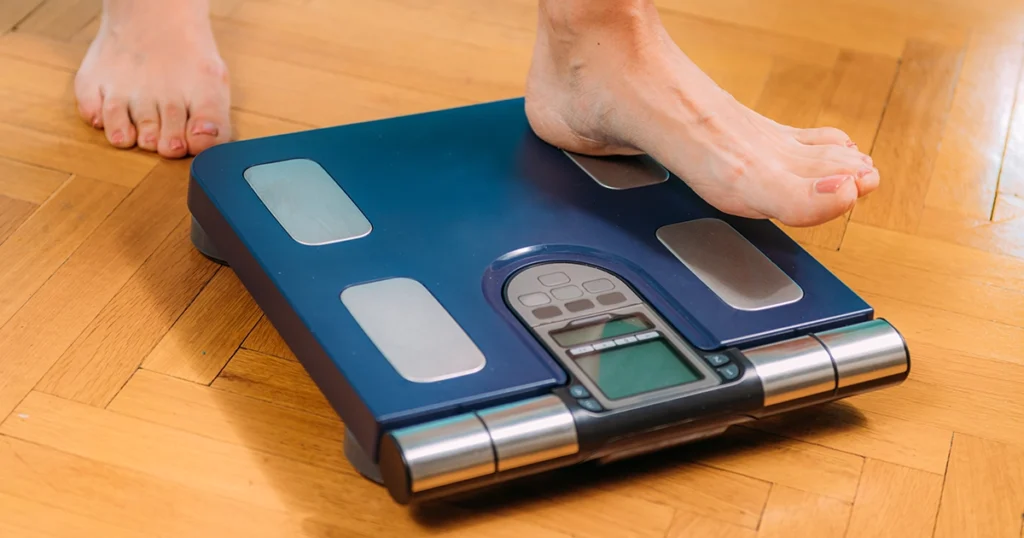 Person standing barefoot on a digital body fat scale on a wooden floor, representing BMI measurement and accurate body composition tracking for people with limb loss or amputation