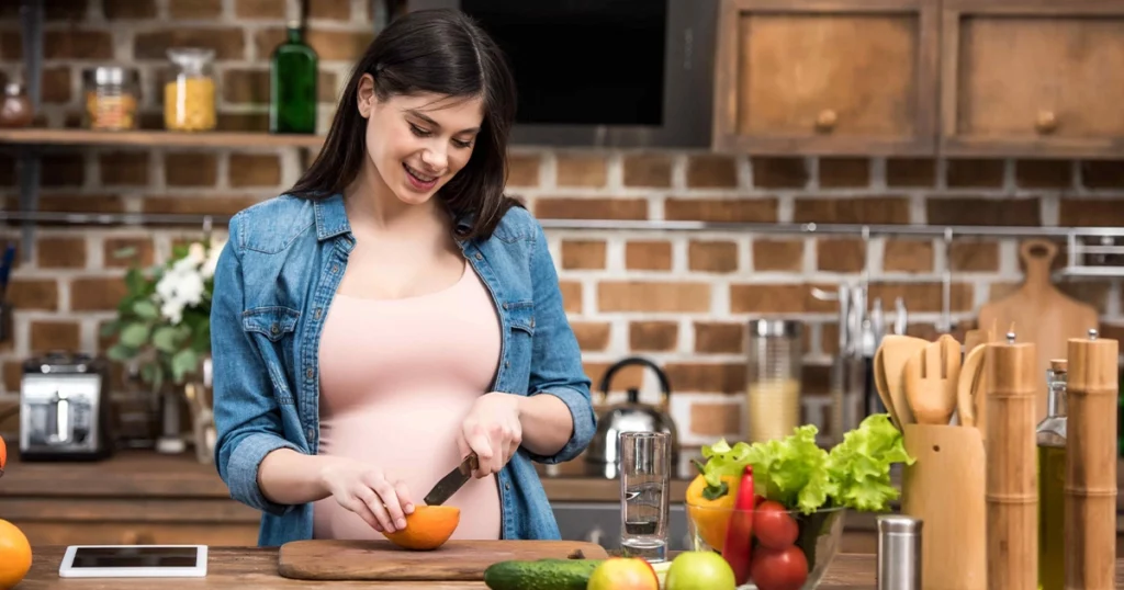 Smiling pregnant woman cutting an orange on a wooden chopping board in a rustic kitchen surrounded by fresh fruits and vegetables, representing healthy nutrition during pregnancy