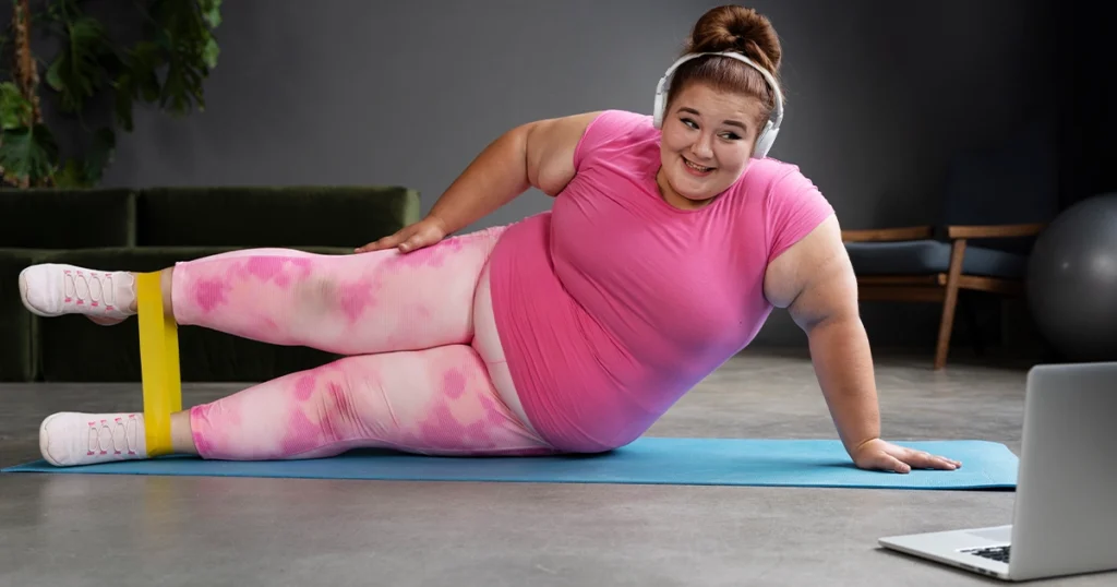 Plus-size woman in pink activewear doing a resistance band leg exercise on a yoga mat while following an online workout, representing fitness and BMI body age calculation