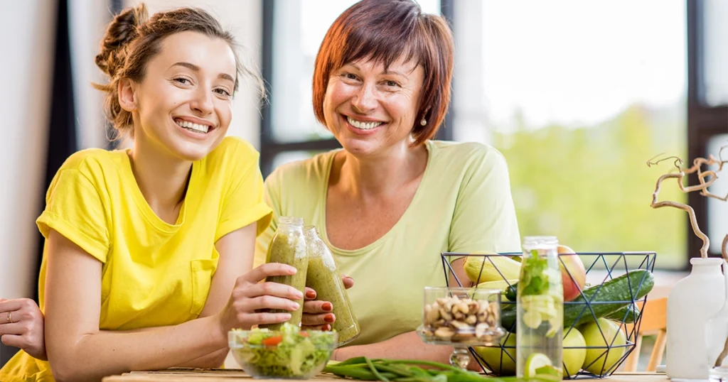 A young woman and an older woman smiling together at a table with healthy food including a green smoothie, fresh fruits, vegetables, and nuts, representing how to improve your body age naturally