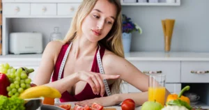 Fit woman with a measuring tape around her neck sitting at a kitchen table surrounded by fresh fruits and vegetables, representing a liquid diet for weight loss